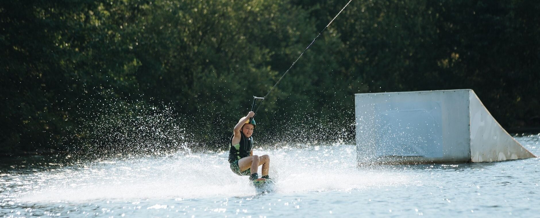Kind fährt beim Wakeboarden an einer Rampe über einen Binnensee
